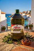 A large 1-liter bottle of Alban cold pressed olive oil stands on a stone ledge outdoors, with a bowl of olives and olive branches in front and a coastal town and blue sea in the background.